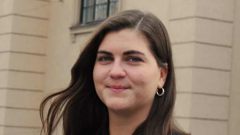 En person med brunt hår klädd i en mörkblå skjorta står framför en gul byggnad.