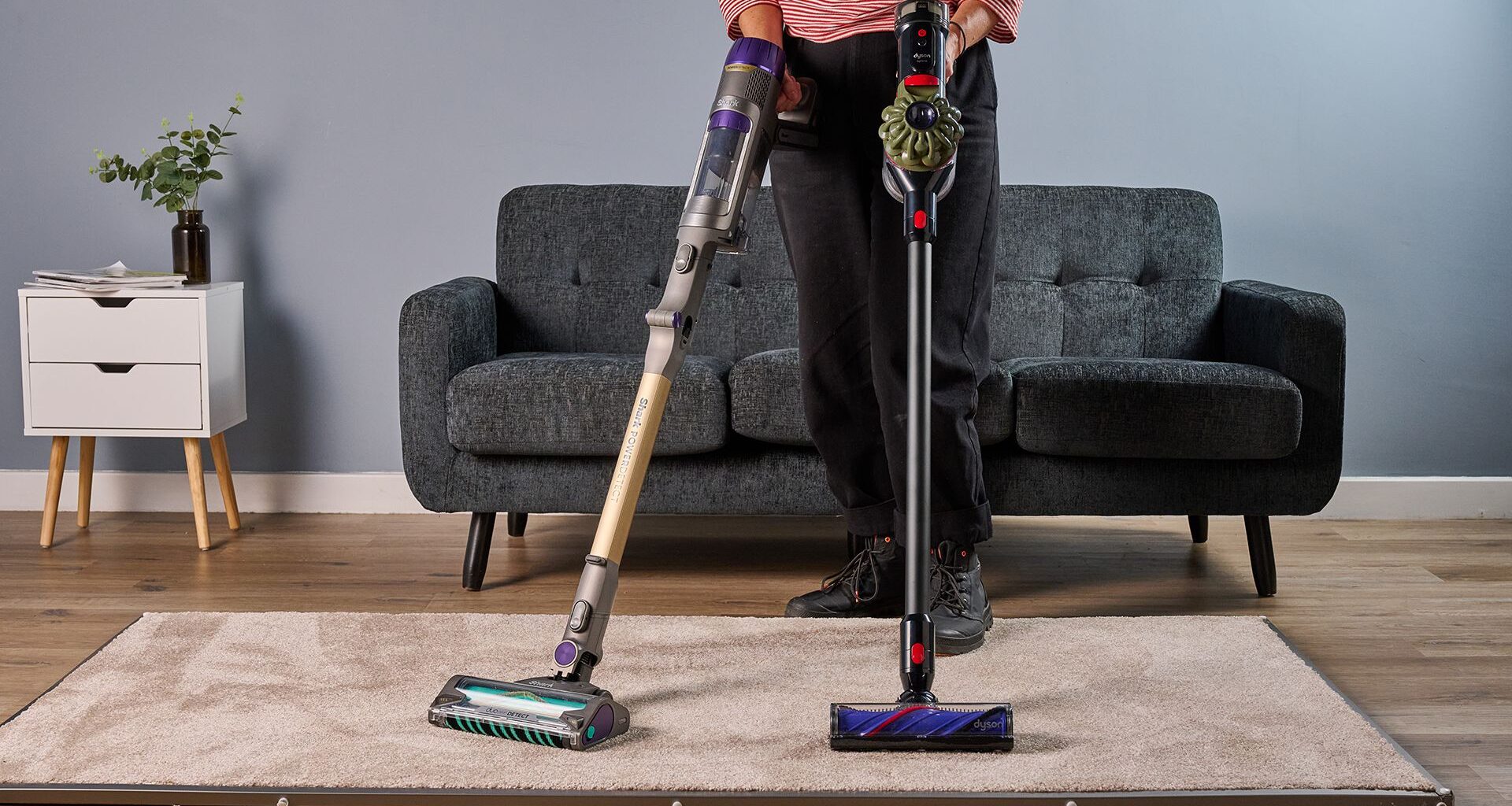 Ruth Hamilton håller sladdlösa dammsugare av typen Shark PowerDetect Cordless och Dyson V8 Cyclone, med en soffa i bakgrunden.