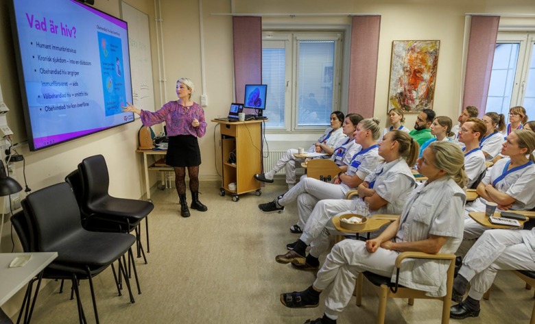 Sjuksköterskan Alexandra Arrhén står och tittar på en stor bildskärm framför ett tjugotal personer iförda arbetskläder som sitter ner och lyssnar på Alexandras föreläsning.