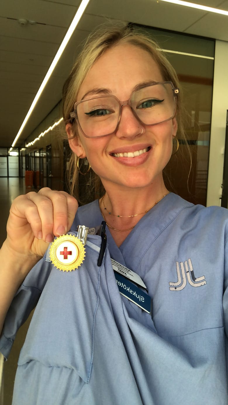 Sjuksköterskan Alexandra Arrhén är iklädd ljusblå arbetskläder och håller stolt fram en rund brosch med ett rött kors i mitten.
