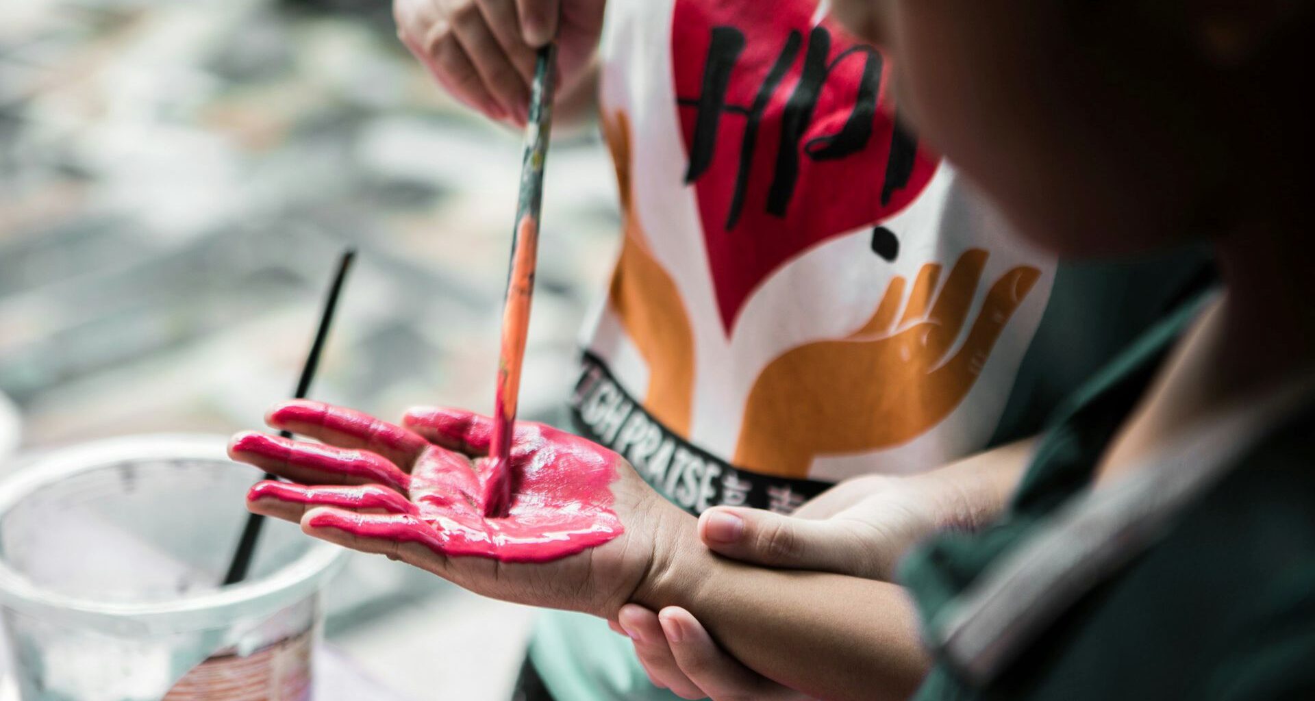 Ett barn målar med rosa färg på ett annat barns handflata, med en pensel.