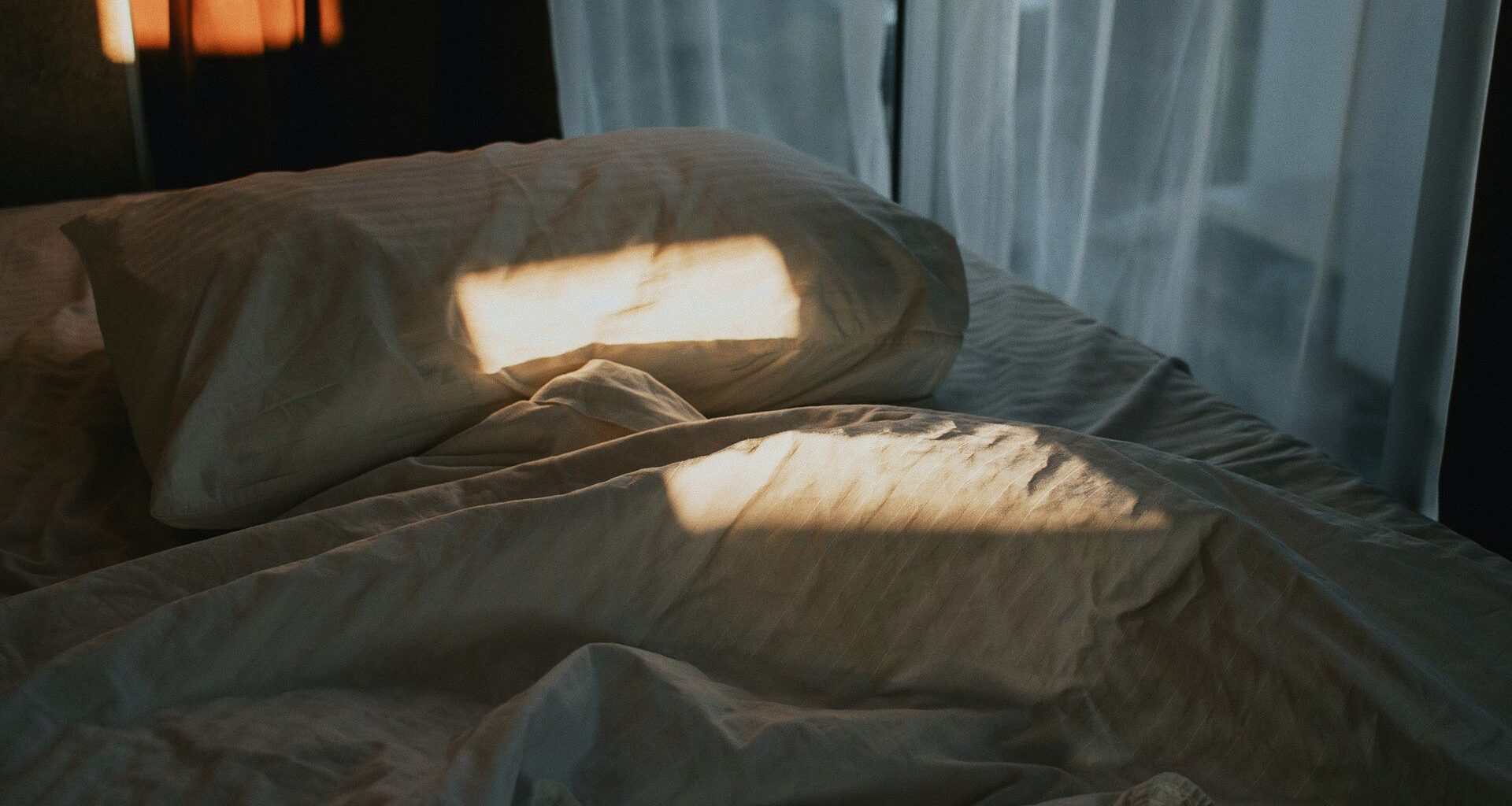 Morgonljus landar fläckvis på en säng med en vit huvudkudde och vitt lakan, gardin bakom.