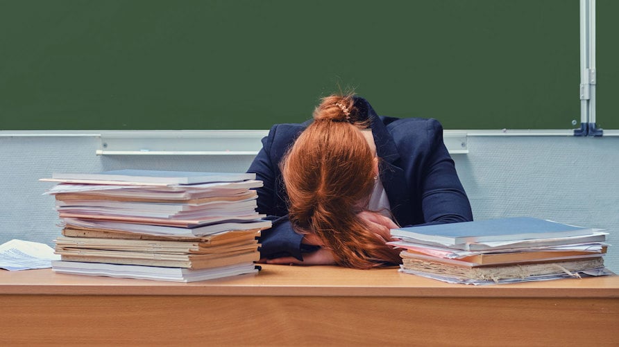En genrebild av en lärare som ligger med ansiktet ned i katedern, buntar av papper framför sig.