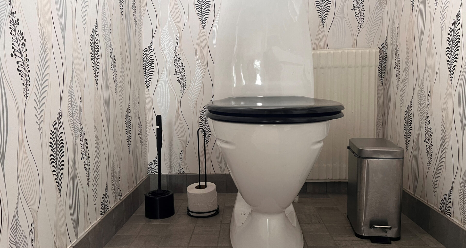 Toalettstol med papperskorg, toaborste och extra toapappersrulle.