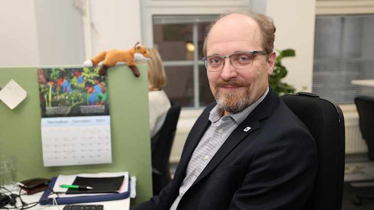 En man i medelåldern sitter vid ett skrivbord i kontorsmiljö, iklädd kavaj. I bakgrunden syns en kalender och en leksaksräv. Bernt Nordman, klimatexpert, WWF.