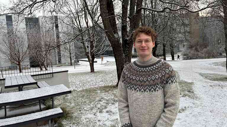 En man står i ylletröja framför ett träd i en snöig park.