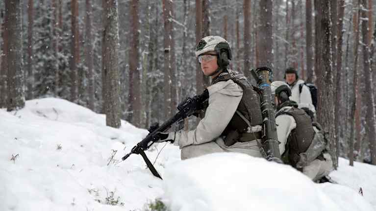 Soldater tränar i skogen under vinterförhållanden, klädda i kamouflage för snömiljöer och utrustade med vapen och kommunikationsutrustning.