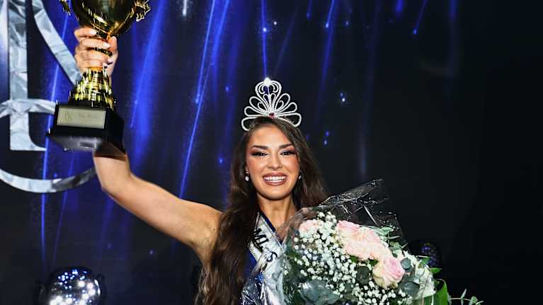 En kvinna med långt brunt hår och krona på huvudet ler och tittar in i kameran. Hon håller i en stor bukett blommor och en pokal.