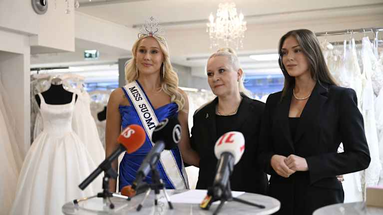 En äldre kvinna står mellan en blond kvinna med Miss Finland-band över axel och brunett utan band och tiara.