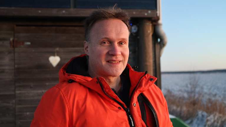 En leende man iförd orange vinterjacka och fotograferad utomhus i gryningsljus en vintermorgon.