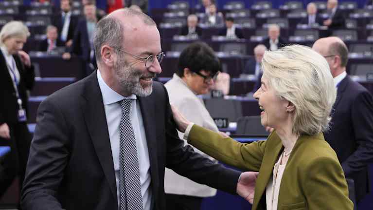 En man med grått skägg och en blond kvinna ler mot varandra i en plenisal.