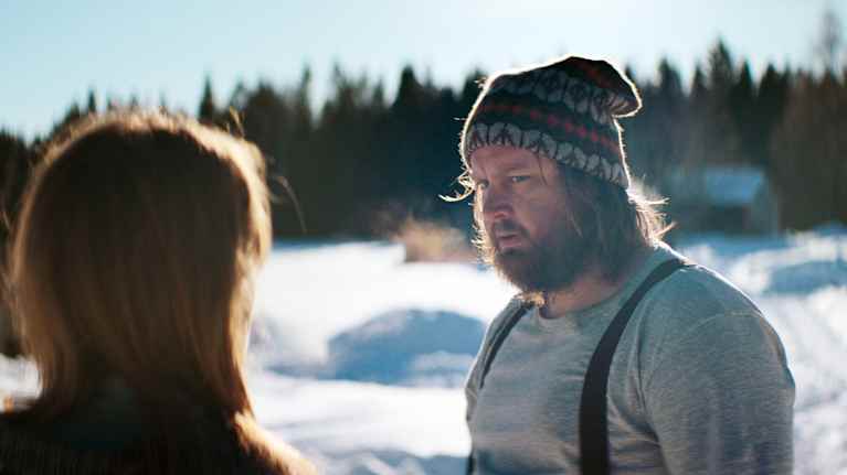 En man i vintermössa och hängselbyxor står i solskenet och tittar på en kvinna som står med ryggen mot kameran.