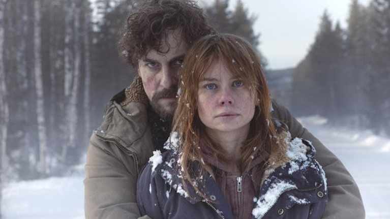 En man med intensiv blick håller om en kvinna med skrämd blick stående på en snöig väg.