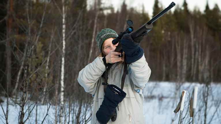 En kvinna i vita vinterkläder riktar ett gevär upp mot himlen.