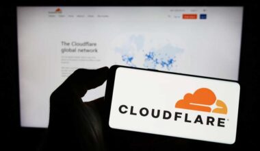 A photograph of a person holding a smartphone with the Cloudflare logo displayed. Behind the phone is a blurred monitor displaying the Cloudflare website.,