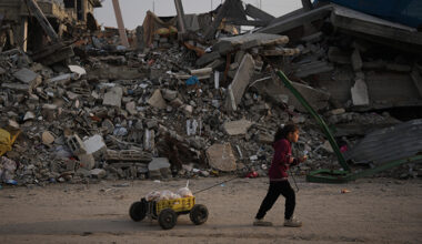 Ett barn går med en vagn. Bakom henne syns förstörda hus.