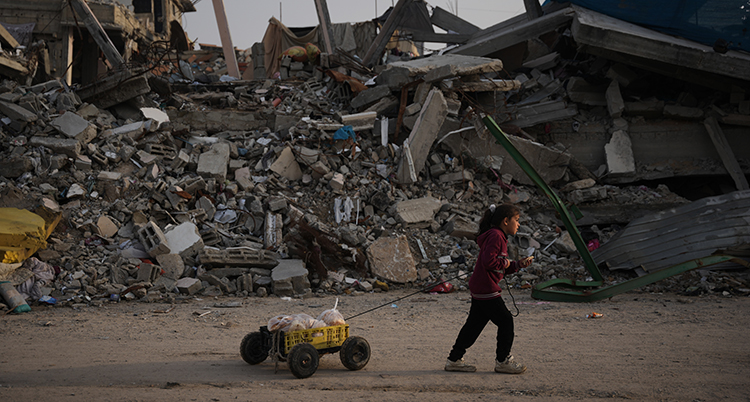 Ett barn går med en vagn. Bakom henne syns förstörda hus.