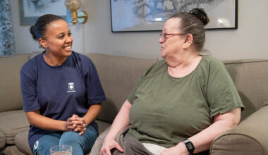 Kvinnlig hemtjänstpersonal i glatt samspråk med kvinnlig äldre brukare i vardagsrumssoffa.