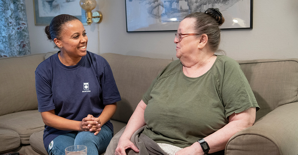 Kvinnlig hemtjänstpersonal i glatt samspråk med kvinnlig äldre brukare i vardagsrumssoffa.