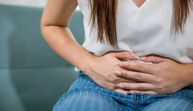 Ung kvinna i vit tröja håller händerna över magen.