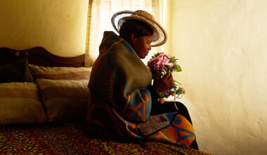 Hon sitter på sängen i ett mörkt rum. Hon har en bukett med blommor i famnen.