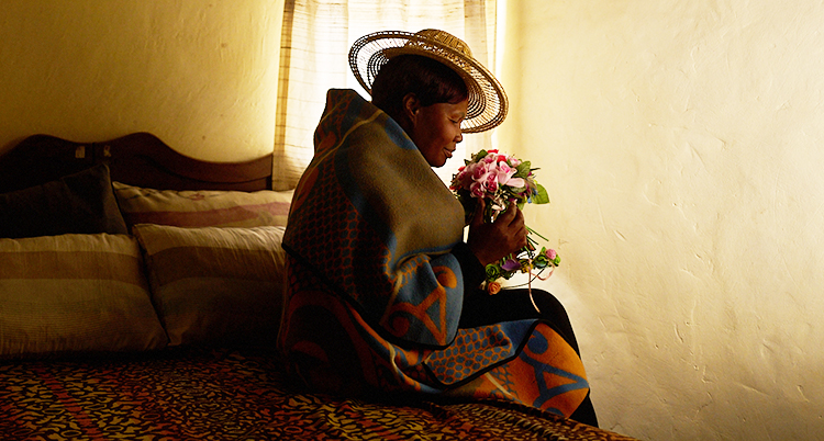 Hon sitter på sängen i ett mörkt rum. Hon har en bukett med blommor i famnen.