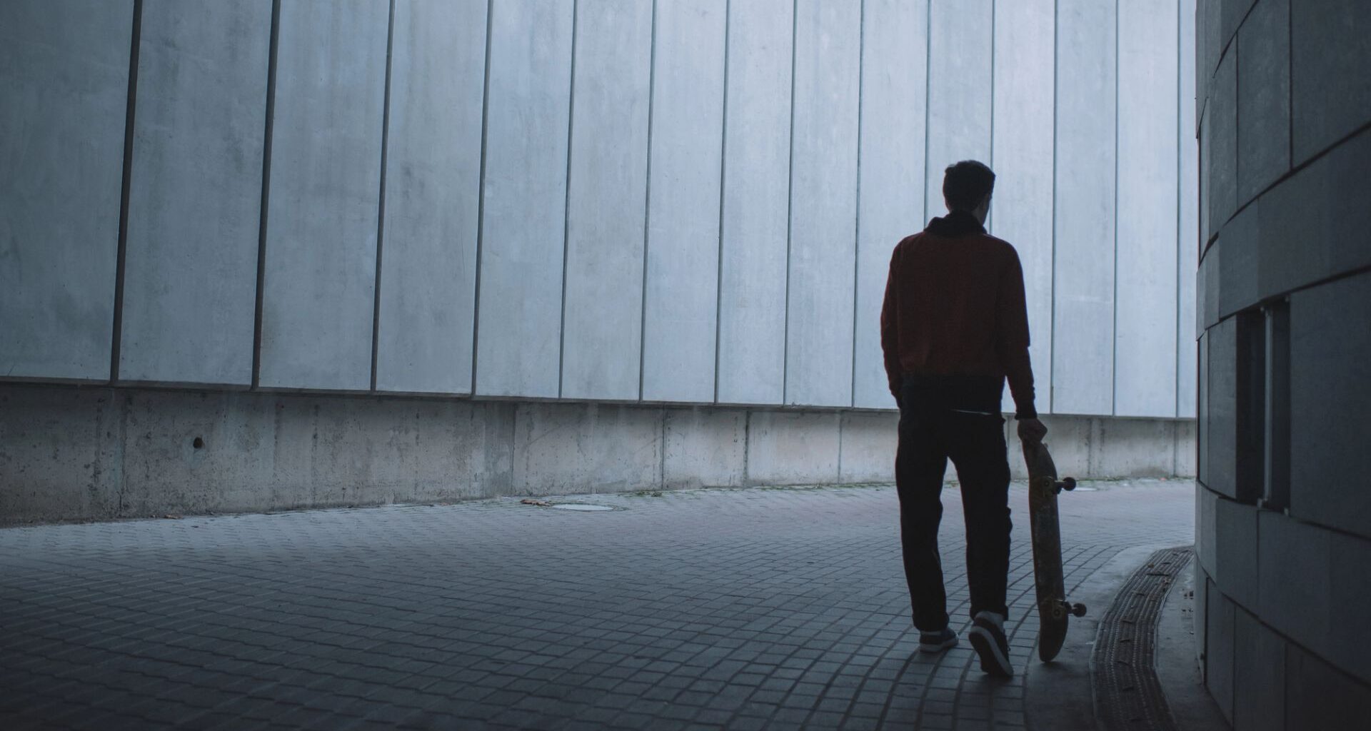 Man i svart siluett står med skateboard i handen i betongtunnel.