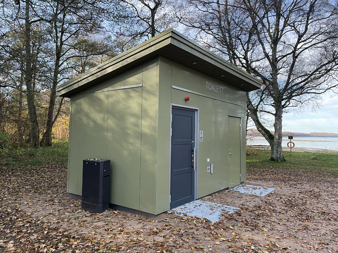 En ljusgrön byggnad i trä vid badplatsen i Gamla Bo med en tillgänglighetsanpassad toalett.