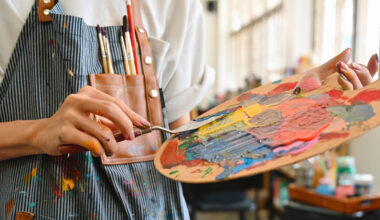 Foto av en person med ett kladdigt förkläde med penslar i bröstfickan. Personen håller i en palett med en massa färger.som håller i en palett med en massa färg