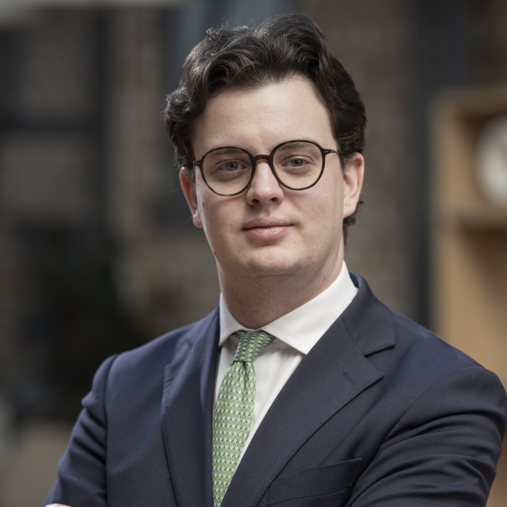 En ung man med kort mörkt hår och glasögon bär en marinblå kostym, vit skjorta och grön slips. Han står inomhus med armarna i kors och tittar in i kameran. Bakgrunden är suddig med hyllor och en tegelvägg som syns.