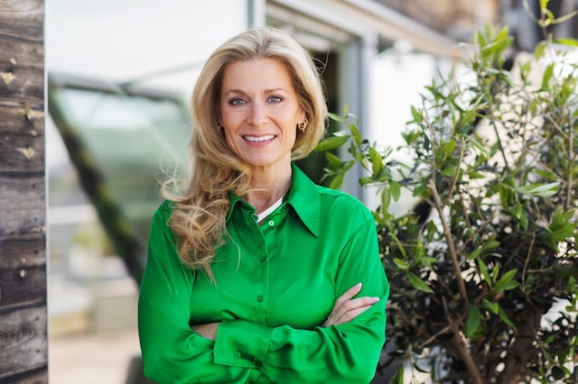 En kvinna med långt blont hår och grön blus står utomhus med armarna i kors och ler framför växter och en byggnad.