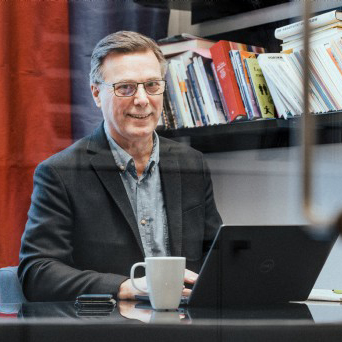 En man med glasögon och kavaj sitter vid ett skrivbord med en bärbar dator och en vit mugg. Böcker ligger staplade på en hylla i bakgrunden. Han tittar in i kameran och ler.