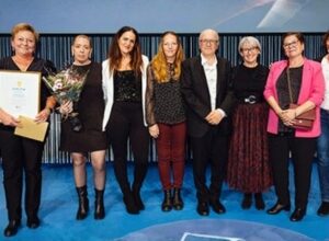 Nio personer står på rad på en scen. De håller i blommor och diplom.