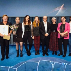 Nio personer står på rad på en scen. De håller i blommor och diplom.