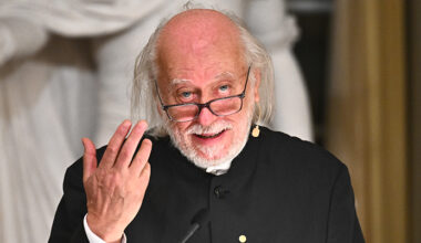 Han tittar in i kameran. Han har glasögon och håller upp ena handen.