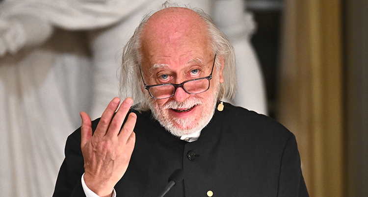 Han tittar in i kameran. Han har glasögon och håller upp ena handen.