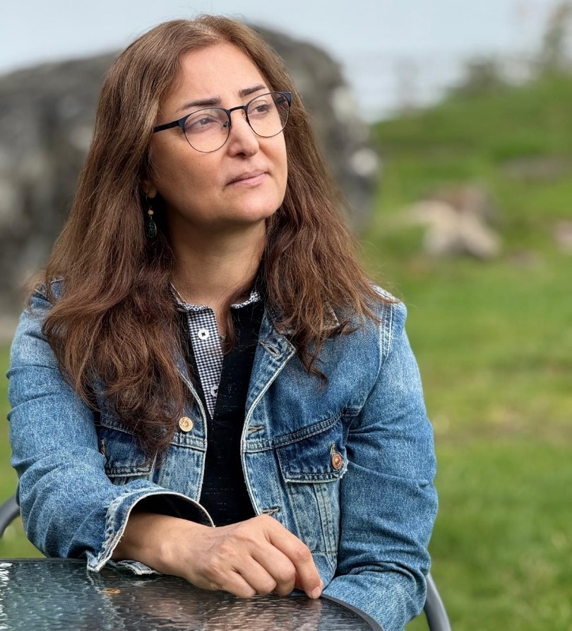 En kvinna med långt brunt hår och glasögon sitter vid ett glasbord utomhus, klädd i jeansjacka. Hon tittar bort åt sidan med ett eftertänksamt uttryck. I bakgrunden finns grönska och en stor sten.