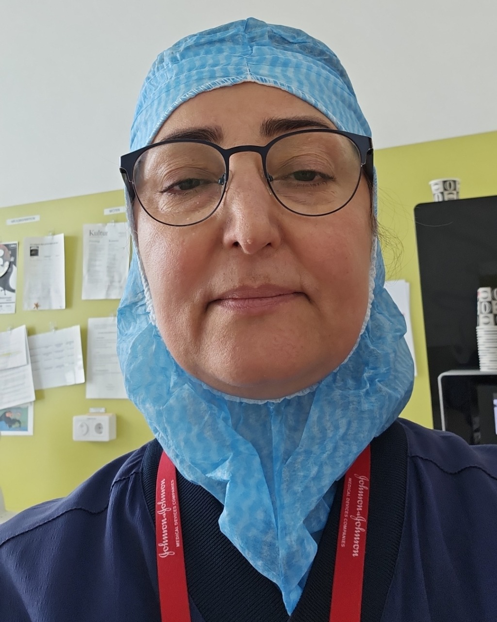 En person med glasögon och en blå engångsmössa med bouffant står inomhus. Mot en gul vägg med papper, anteckningar och en svart tavla tittar de direkt in i kameran - kanske reflekterar de över ett spårbyte utvisas.