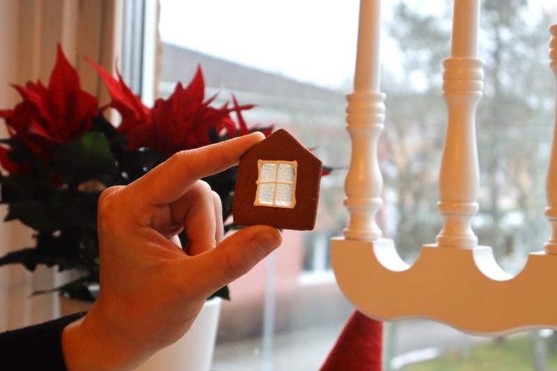 En hand håller en liten husformad kaka med ett genomskinligt fönster framför ett fönster; en julstjärna och en vit ljusstake finns i bakgrunden.