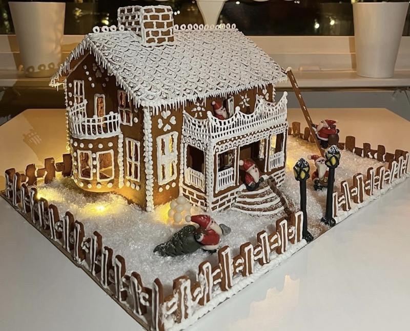 Pepparkakshus dekorerat med vit glasyr, omgivet av ett staket, med små tomtefigurer som klättrar upp för en stege och är utspridda på gården. Scenen är belyst från insidan av huset.