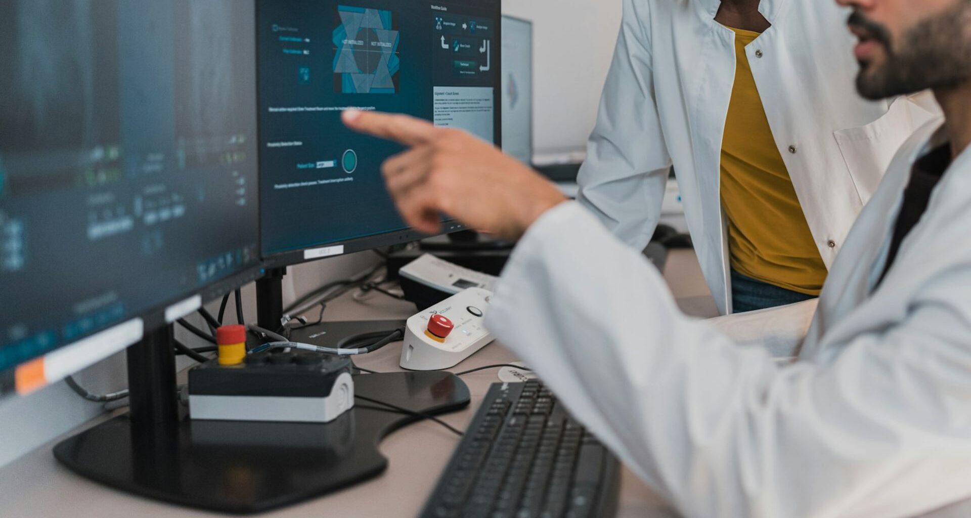 En person i vit rock, vid skrivbord, pekar mot en datorskarm. Annan person i vit rock i bakgrunden.