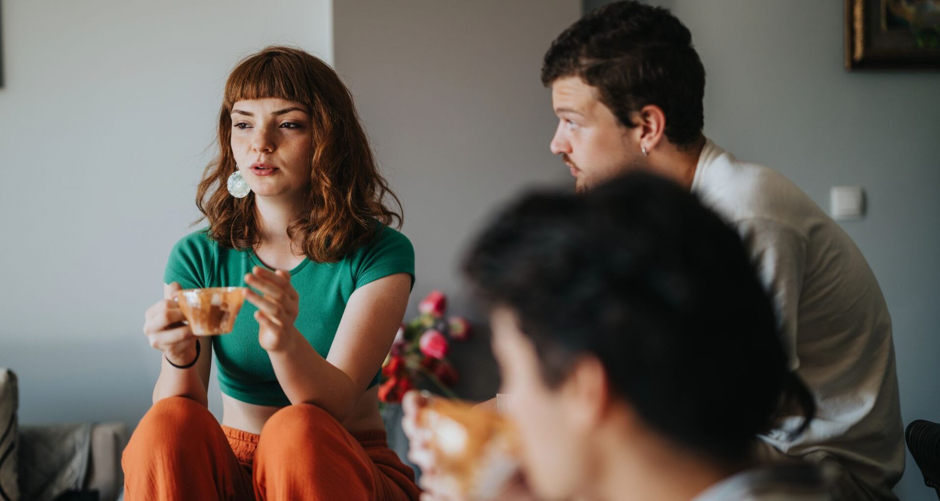 Tre unga personer sitter i hemmiljö och pratar. Ung kvinna med långt hår och tekopp säger något.