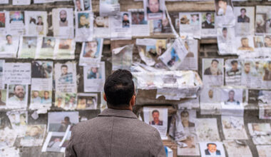 Mannan står med ryggen mot kameran. Väggen är fylld av papper och bilder på personer.