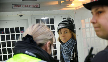 Hon har en mössa och sjal på sig. Hon står bakom två poliser.