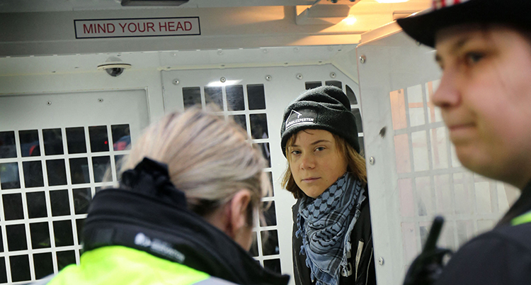 Hon har en mössa och sjal på sig. Hon står bakom två poliser.