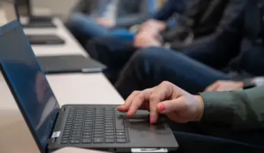 Närbild på laptops på ett bord. Vid bordet sitter tre personer.