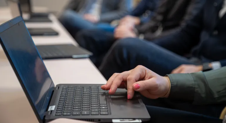 Närbild på laptops på ett bord. Vid bordet sitter tre personer.