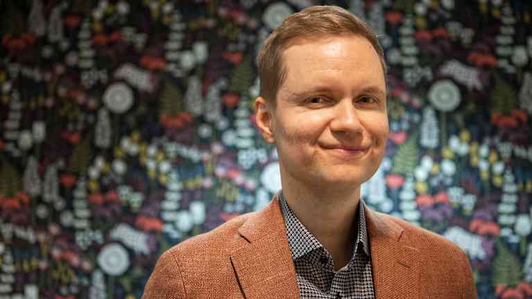 Bilden visar en person iklädd en brun kavaj och en rutig skjorta, stående framför en färgglad, mönstrad bakgrund.