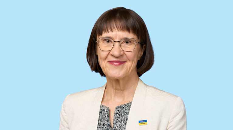 En kvinna i vit kavaj och spräcklig grå skjorta framför en blå bakgrund.