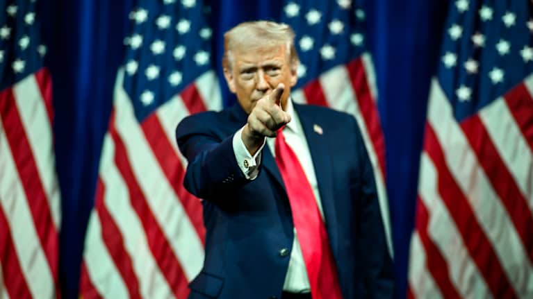 Donald Trump står framför en rad amerikanska flaggor och pekar mot publiken med höger pekvinger.